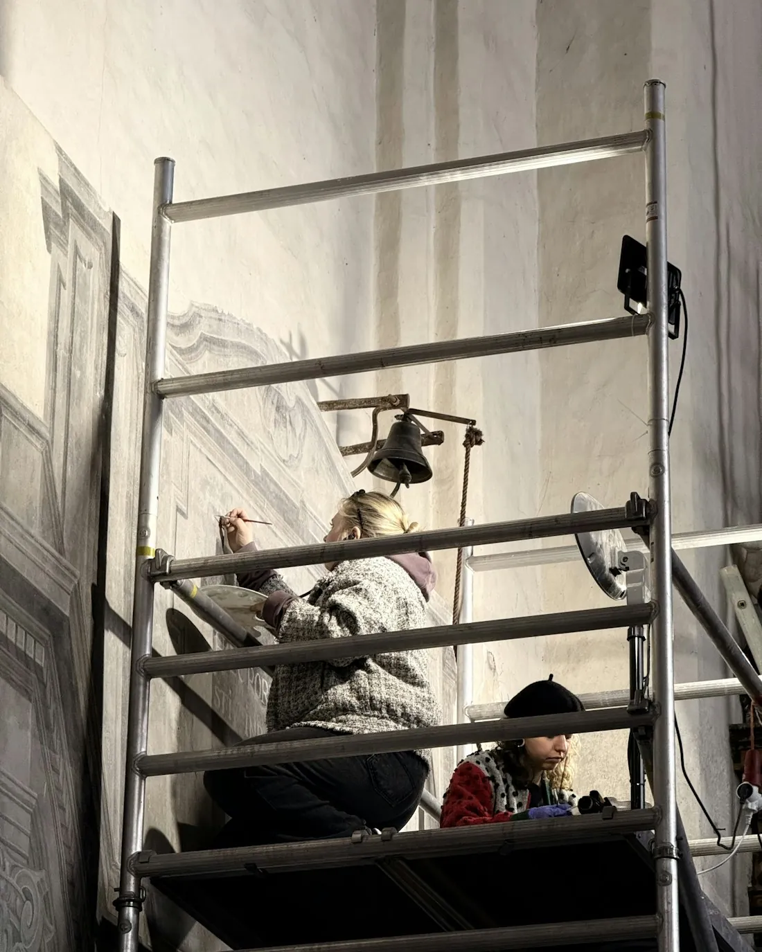 Dos personas pintan un muro enlucido desde un andamio en el interior de un edificio